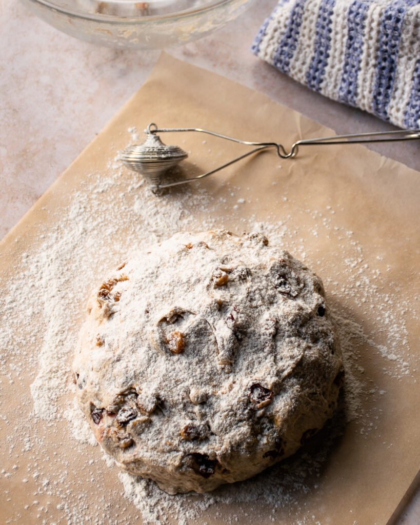 Fruit and nut bread resting on floured parchment paper with a flour duster near by.