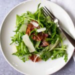 Arugula, Parmesan and Prosciutto salad on a white plate with a fork.