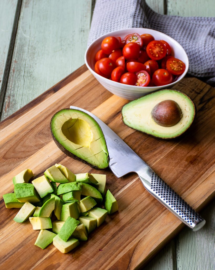 Cutting an avocado on a wooden cutting board with tomatoes in the background.