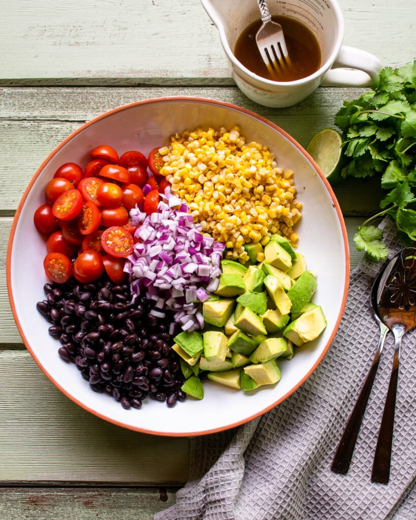 Ingredients for avocado tomato salad in a white bowl with a pitcher of dressing and some cilantro next to it.