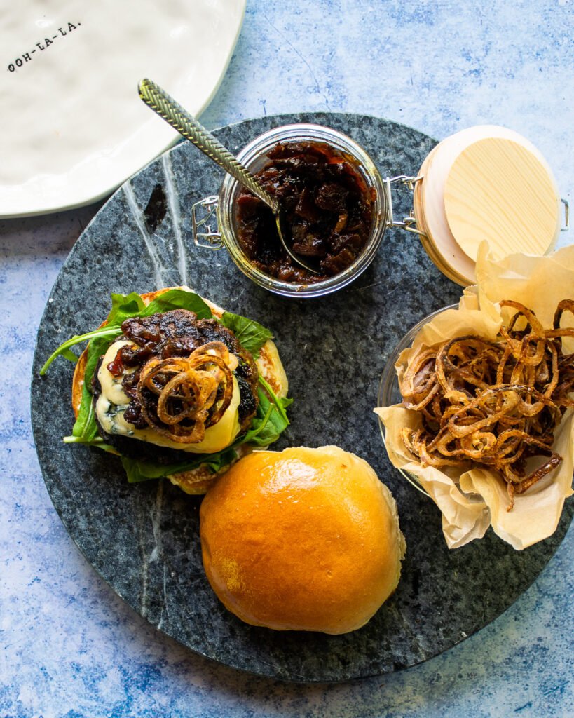 Bacon Jam Blue Burger with Crispy Shallots Looking down on a burger with bacon jam on it next to a jar of bacon jam and some crispy shallots.