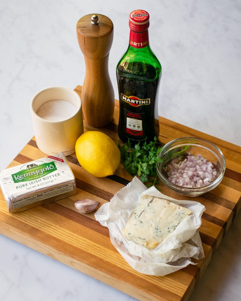 Ingredients on a wooden cutting board - butter, lemon, gorgonzola cheese, shallots, vermouth, salt, pepper, garlic and parsley
