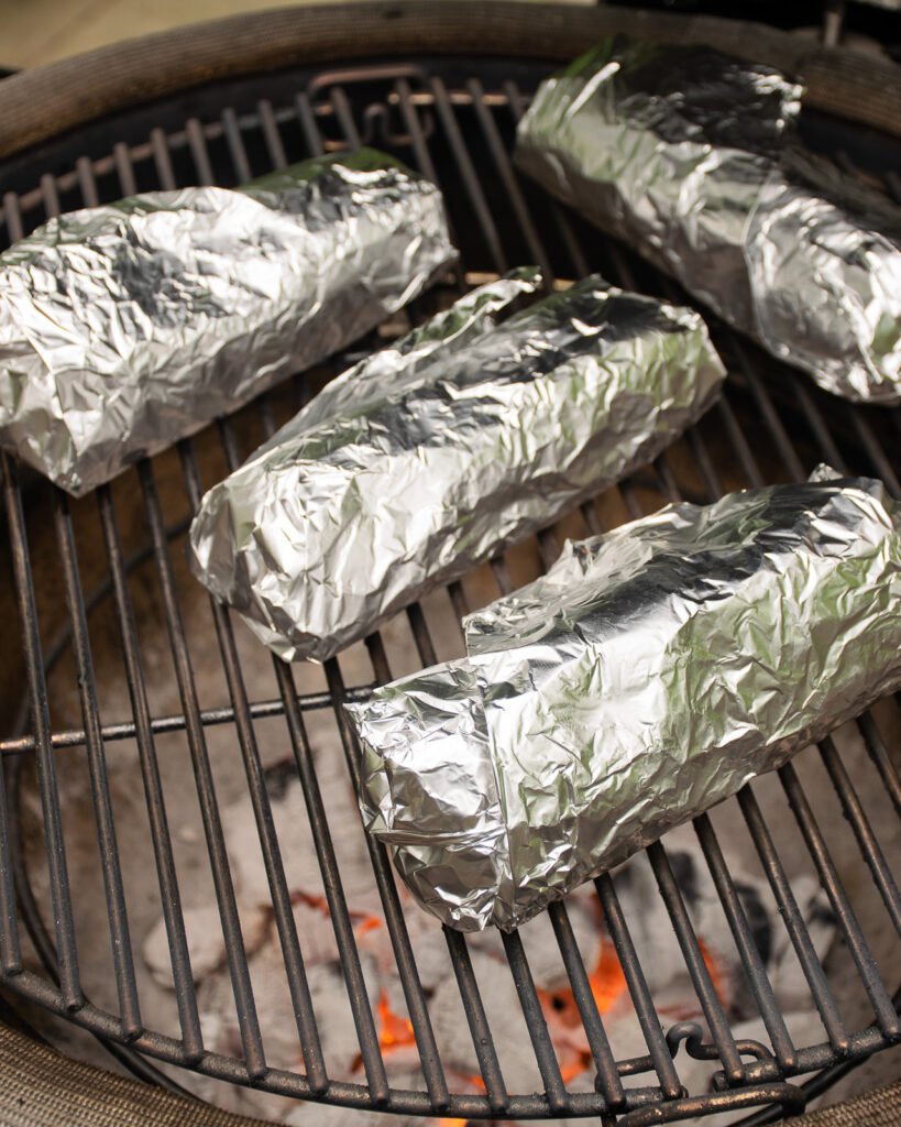 Four roast beef sandwiches wrapped in foil on a bbq grill grate.