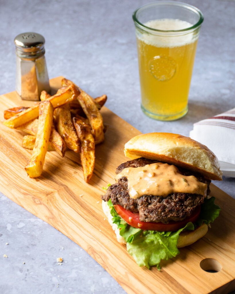 A smash burger and some french fries on a wooden board with a glass of beer and a salt shaker.