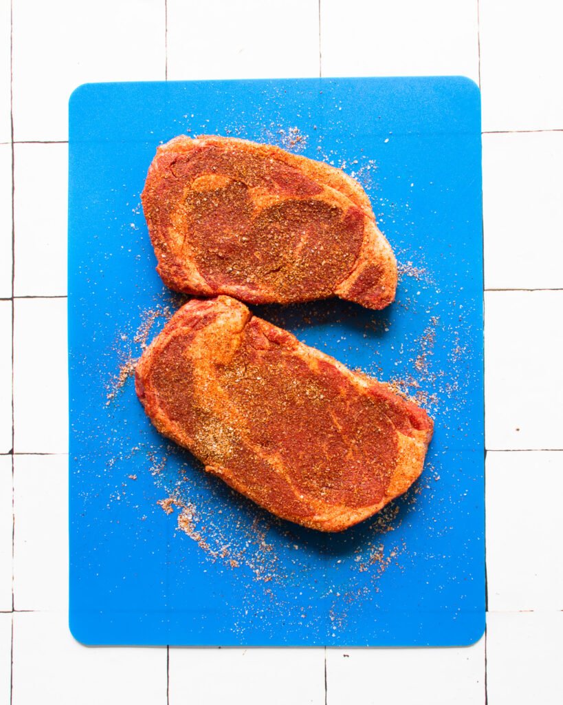Two spice rubbed ribeye steaks on a blue cutting board.