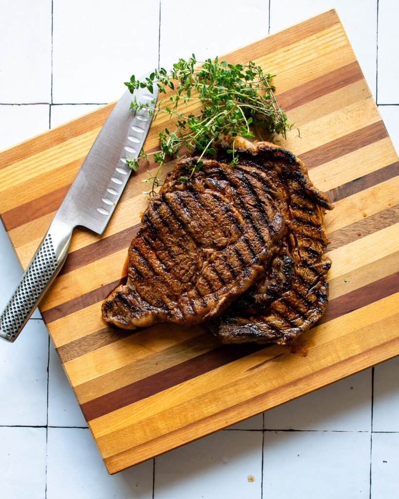 two grilled ribeye steaks on a wooden cutting board with a knife and some fresh thyme.
