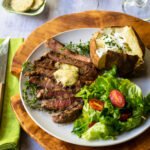 A dinner plate with sliced ribeye steak, a mashed potato and a green salad, along with a glass of wine and salt and pepper shakers.