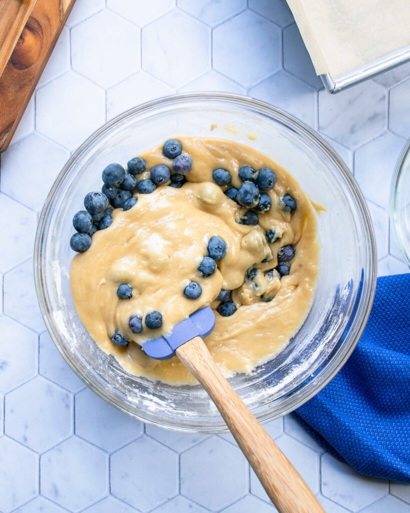 Folding blueberries into a batter in a bowl on a tiled counter.