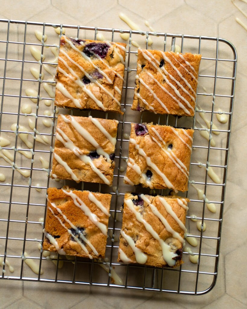 Four blueberry blondies on a cooling rack with white chocolate glaze drizzled on top.