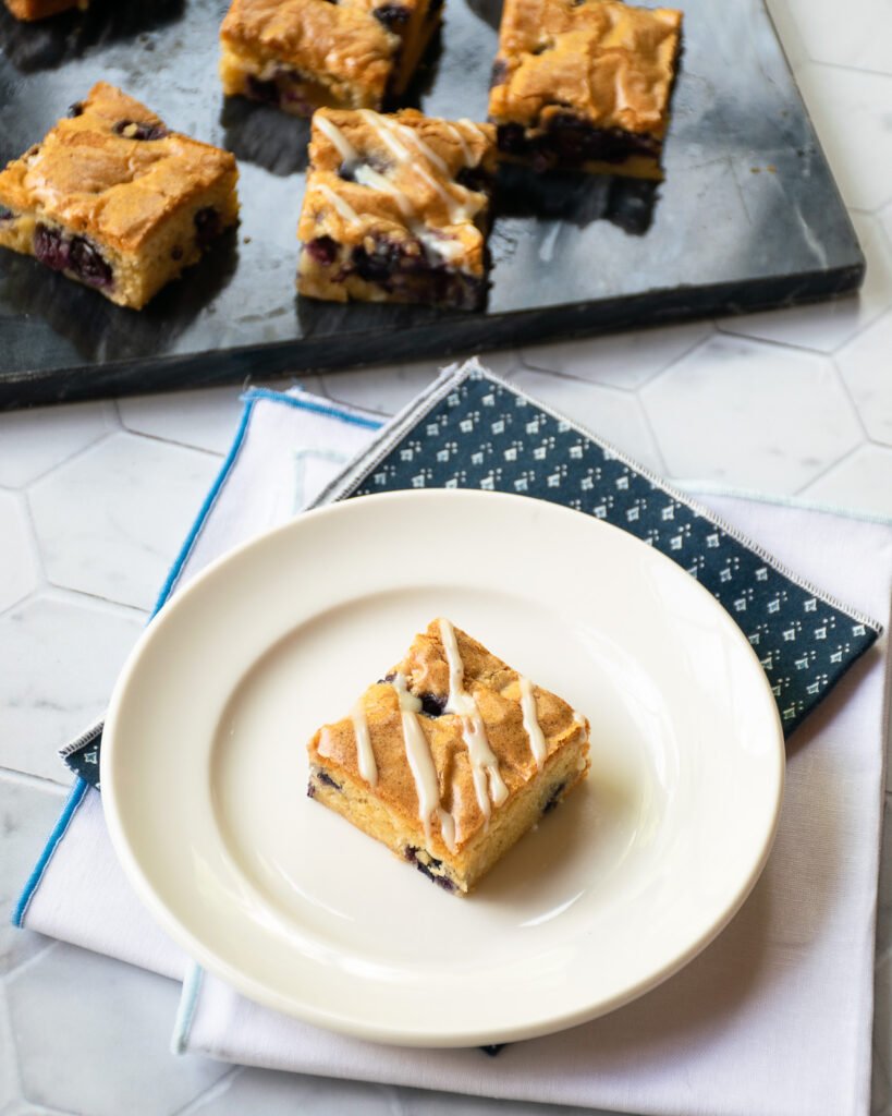 A blueberry blondie on a white plate with more blueberry blondies in the background on black marble.