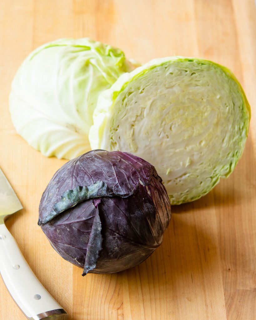 A halved head of green cabbage with a head of red cabbage in front of it and a white handled knife.