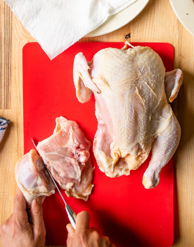 Two hands cutting a chicken leg into drumstick and thigh on a red cutting board, the second step in how to cut up a whole chicken.