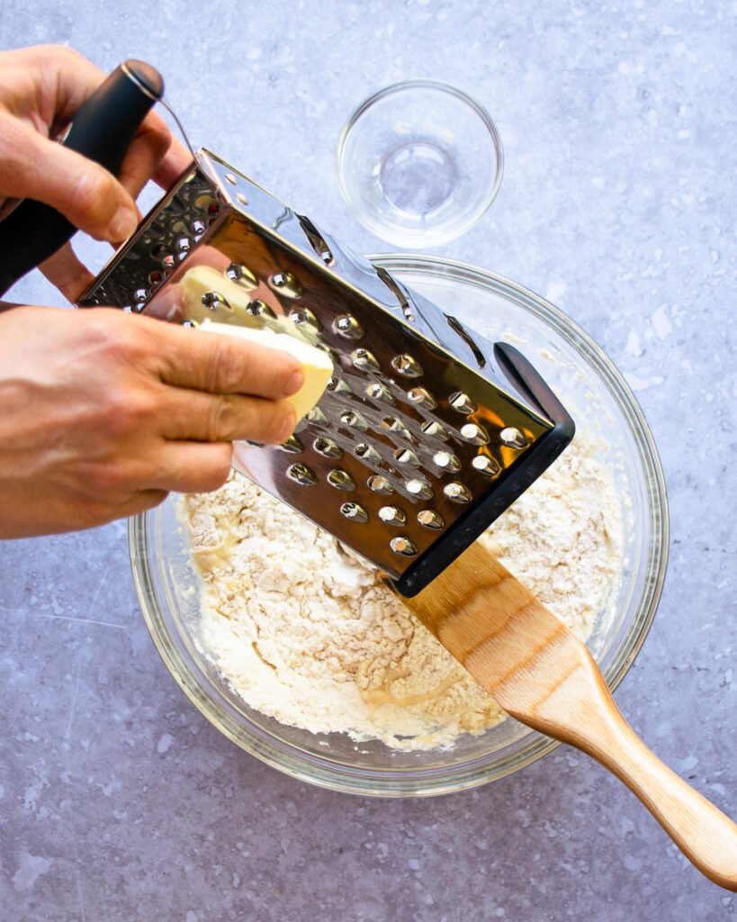 Two hands grating butter into a bowl of flour with a wooden spoon sticking out of it.