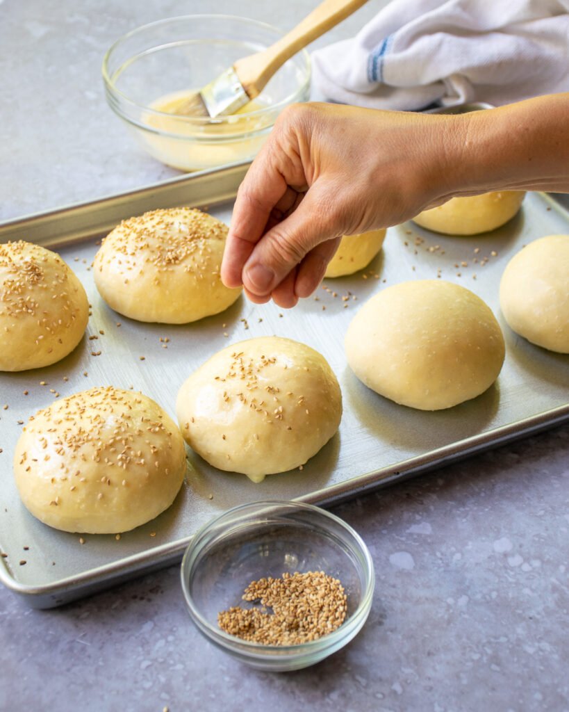 A hand sprinkling sesame seeds on hamburger dough balls.