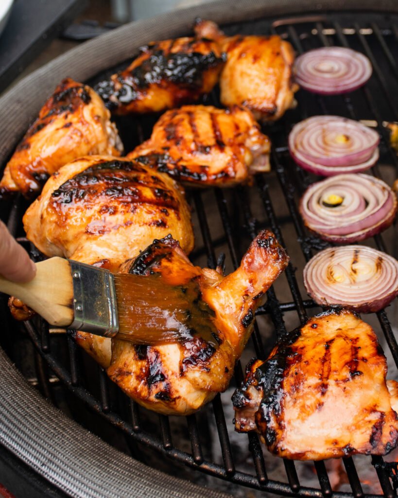 A brush basting Huli Huli chicken on the grill with red onions grilling alongside.