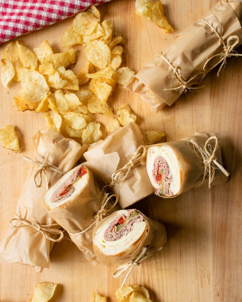 Roast beef sandwiches wrapped in parchment paper on a cutting board with chips scattered around.