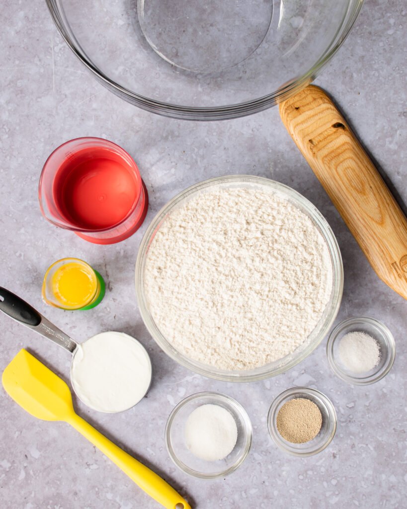 Ingredients on a counter - flour, salt, yeast, sugar, yogurt, melted butter, water - along with a spatula, wooden spoon and glass bowl.