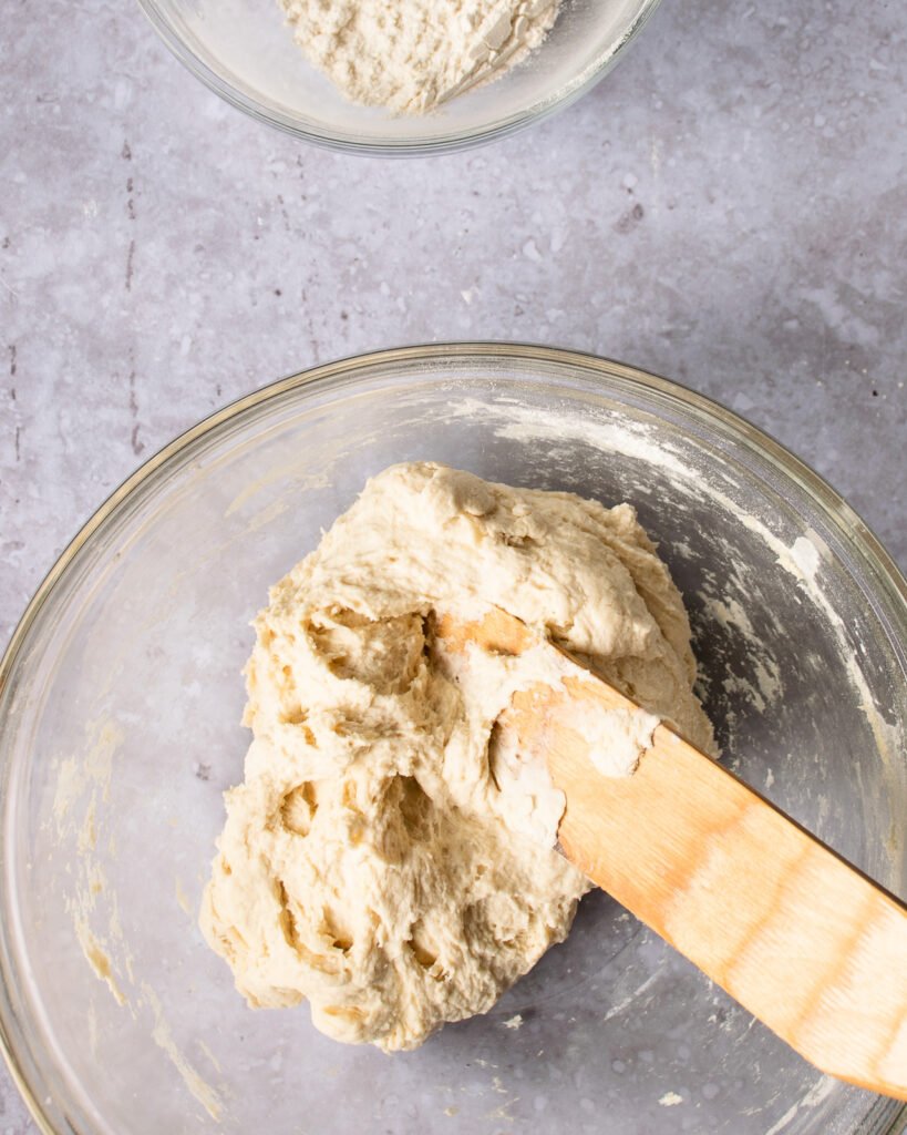 Naan bread dough in a glass bowl with a wooden spoon.