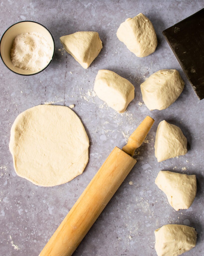 A small bowl of flour, a bench scraper, a rolling pin and small balls of dough with one rolled out into a circle on a counter.