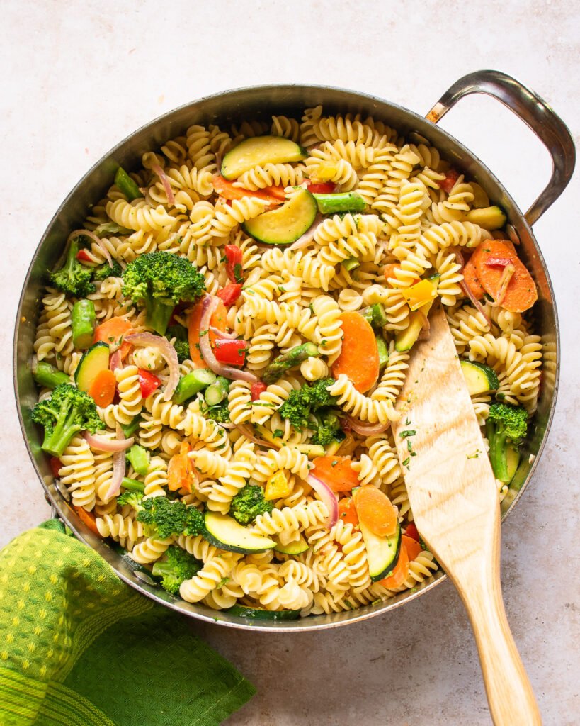 Pasta primavera in a sauté pan with a wooden spoon sticking out.