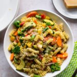 A large family style bowl of pasta primavera with a block of parmesan cheese on a small white plate and a green napkin.