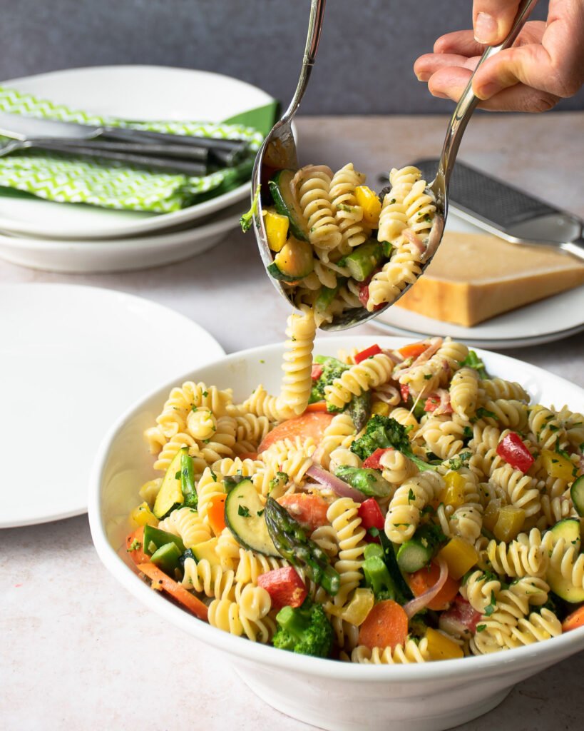 Serving pasta primavera from a large bowl with two serving utensils.