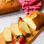 A pound cake sliced on a wooden board with strawberries, a red napkin and a second pound cake in the background.