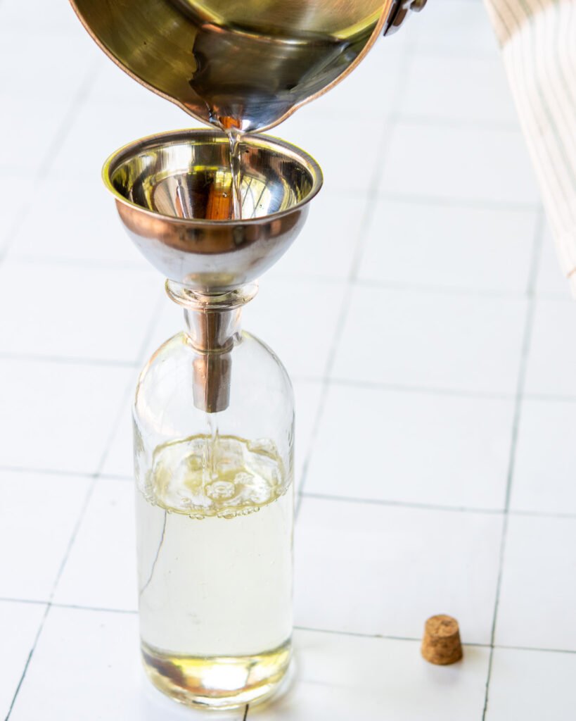 A saucepan pouring simple syrup into a glass bottle through a metal funnel.