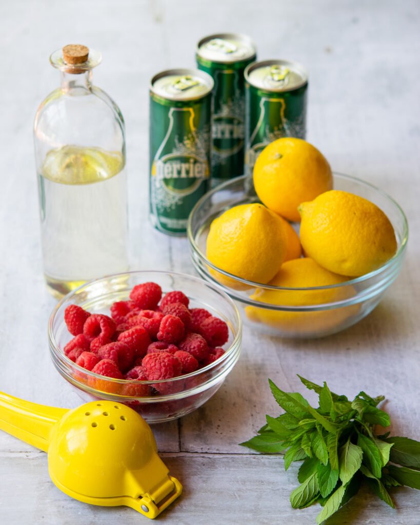 Ingredients on a grey table - sparkling water, lemons, raspberries, simple syrup and mint.