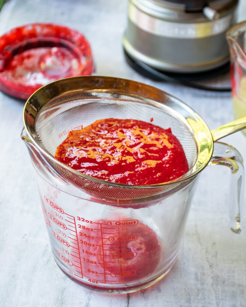 A glass measure and fine strainer on a table with raspberry purée in the strainer.