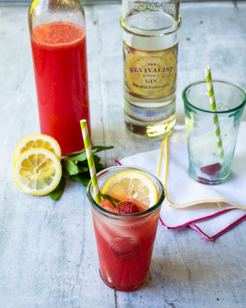 A bottle of sparkling raspberry lemonade next to a bottle of gin with a glass of sparkling lemonade in the foreground.