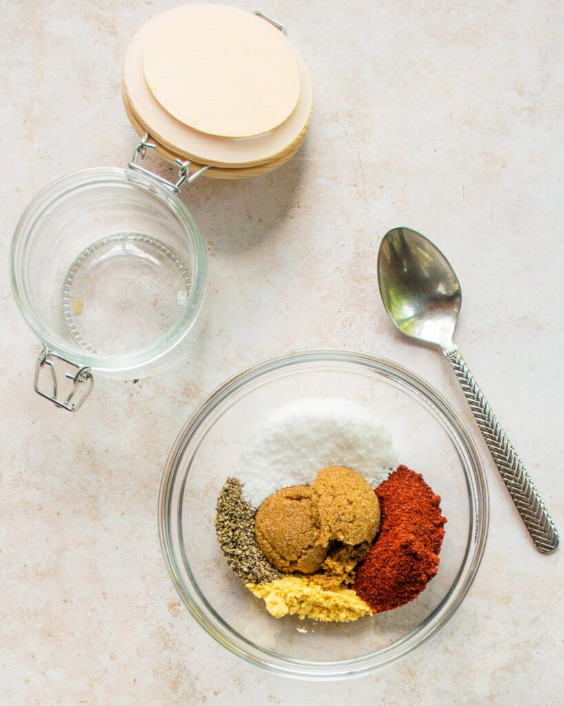Spices mixed in a glass bowl with a spoon and an empty glass jar next to it.