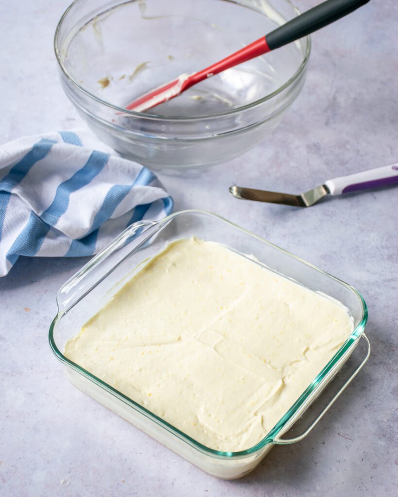 A glass square baking dish with cheese cake filling spread into it, an offset spatula, a blue and white striped towel, an empty glass bowl and a red spatula.