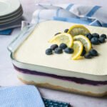 Blueberry lemon delight in a glass square baking dish with a stack of plates in the distance and blue and white towels and napkins.