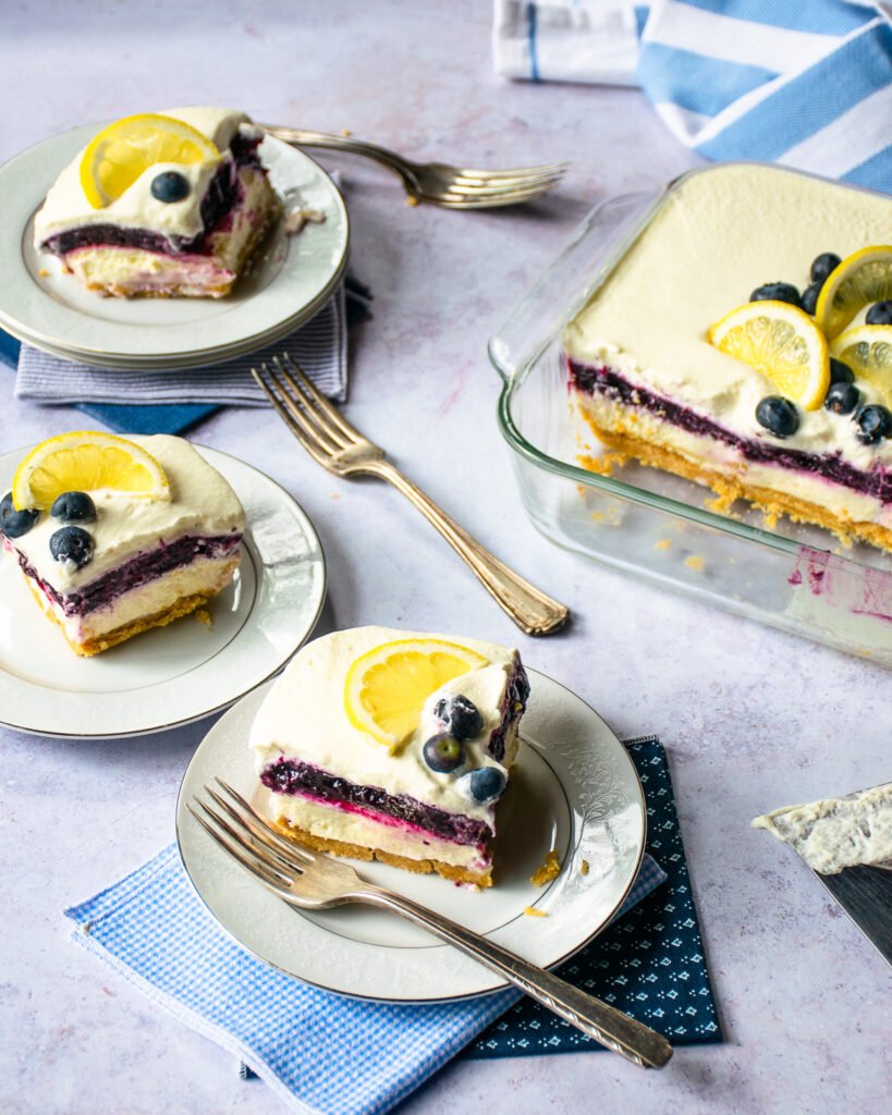 Blueberry Lemon Delight cut into pieces and on small plates with a larger baking dish of the dessert in the background.