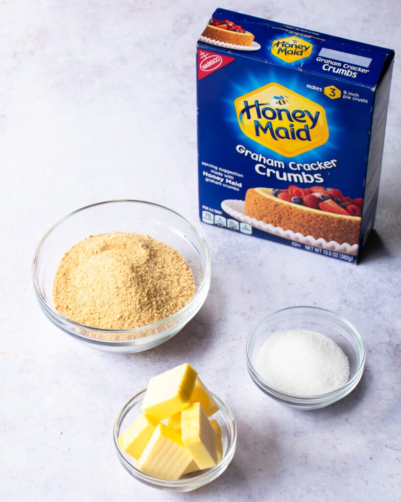 Three ingredients on a countertop - graham cracker crumbs, butter and sugar.