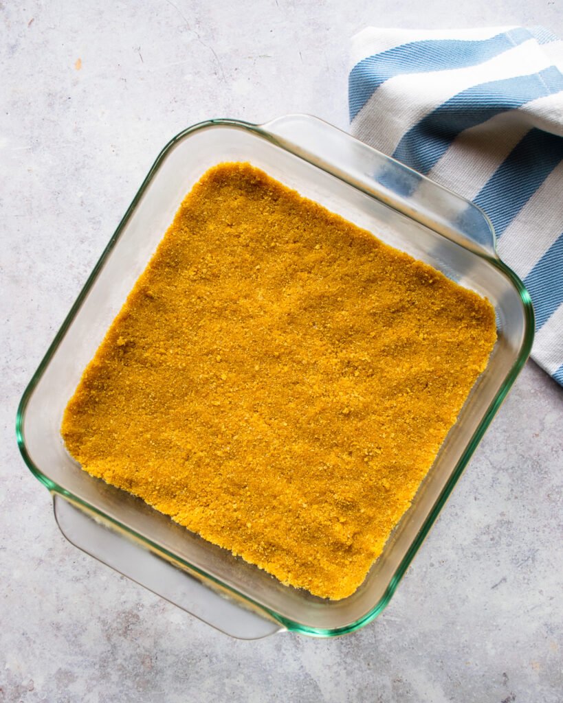 Graham cracker crumb crust pressed into the bottom of a glass square baking dish with a blue and white striped towel.