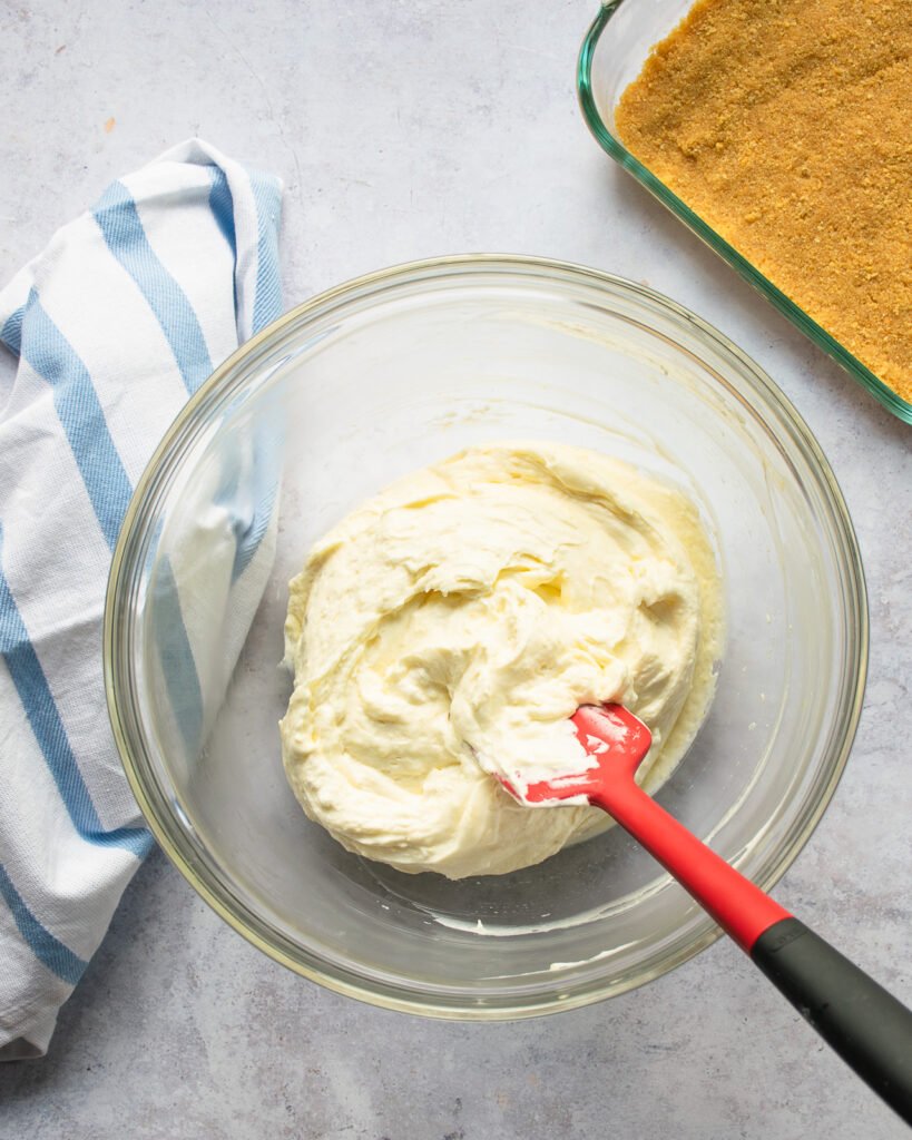 A glass mixing bowl with cream cheese filling and a red spatula, a square glass baking dish with graham cracker crust and a blue and white striped towel.