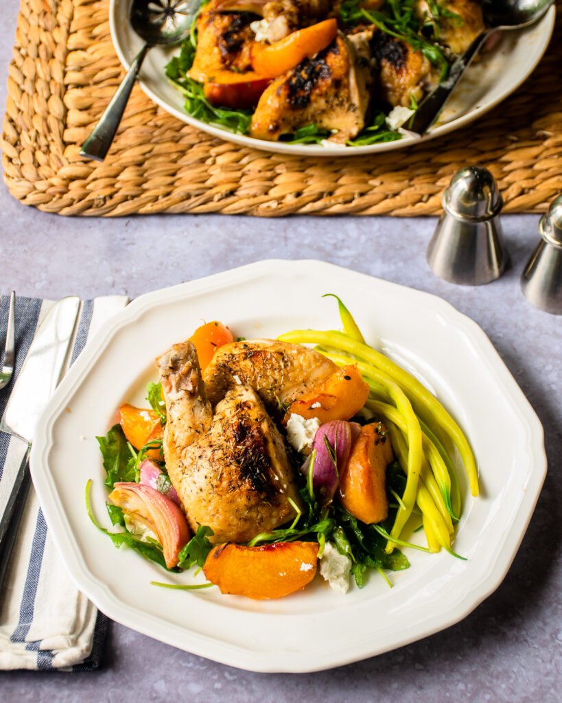 A white dinner plate with chicken and balsamic baked peaches with more on a serving plate near by.