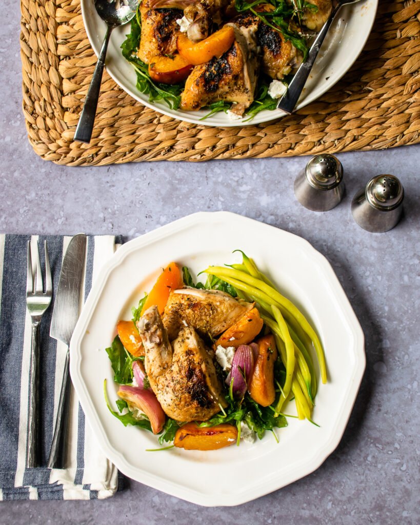 A white dinner plate with chicken and balsamic baked peaches with more on a serving plate near by.