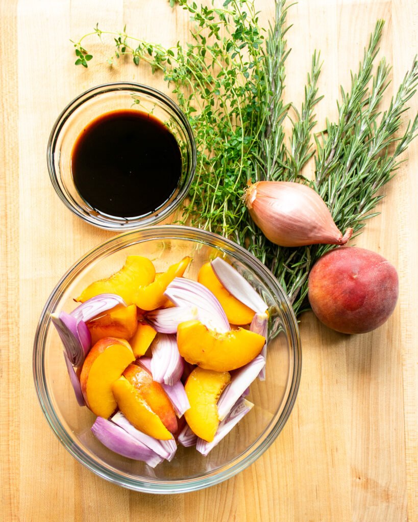 A bowl of peaches and shallots on a cutting board with fresh herbs and a bowl of balsamic vinegar.