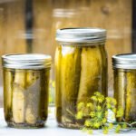 Three jars of pickles on a white counter with a window behind them.