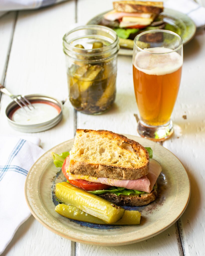 A sandwich on a plate with a dill pickle spear and a glass of beer.