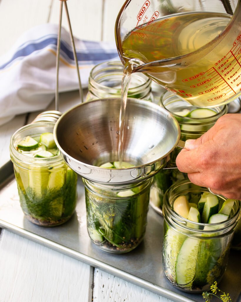 Hands pouring hot pickle brine into jars of cucumbers.