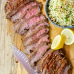 Flat iron steak, sliced on a cutting board with a bowl of couscous and a cut lemon.