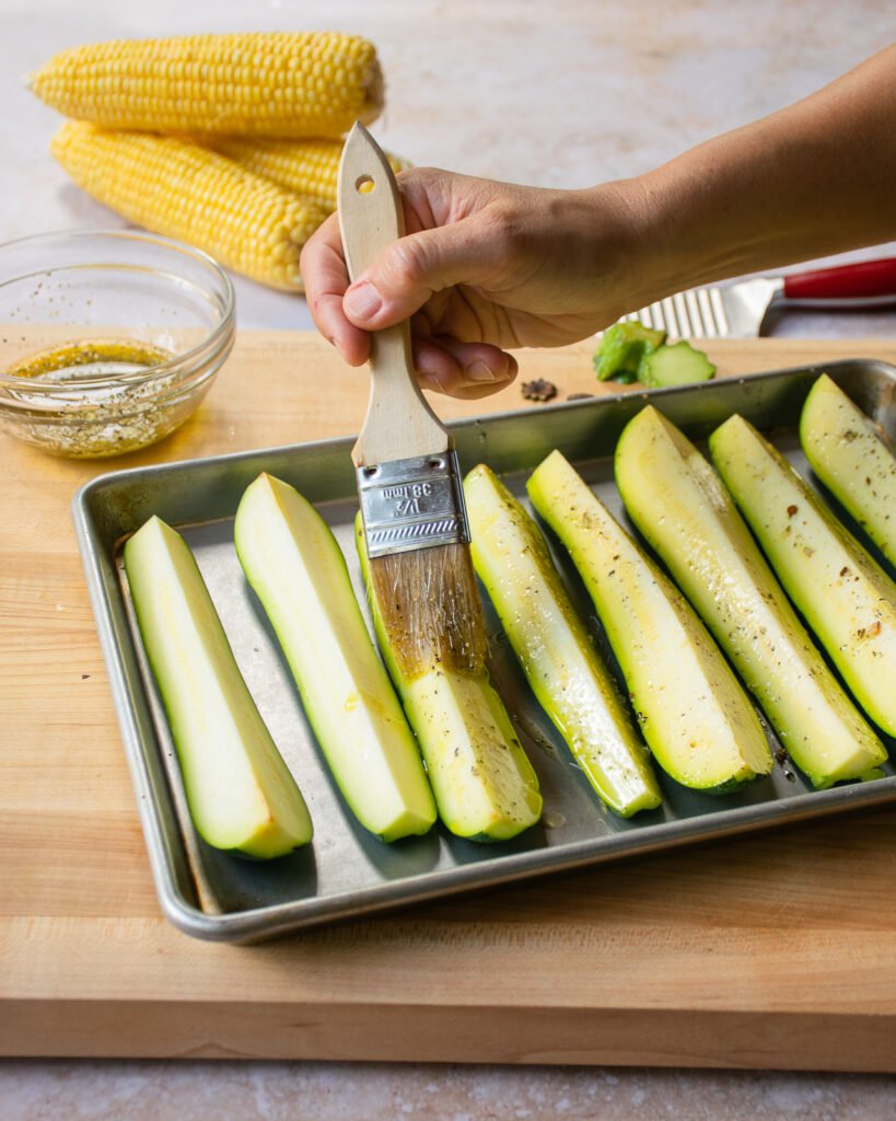 A hand brushing oil on zucchini on a quarter sheet pan with corn in the background.