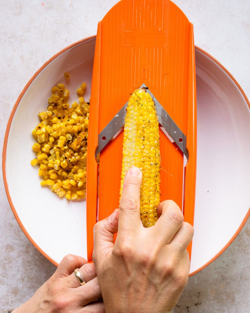 Hands cutting kernels off an ear of corn with an orange mandolin.