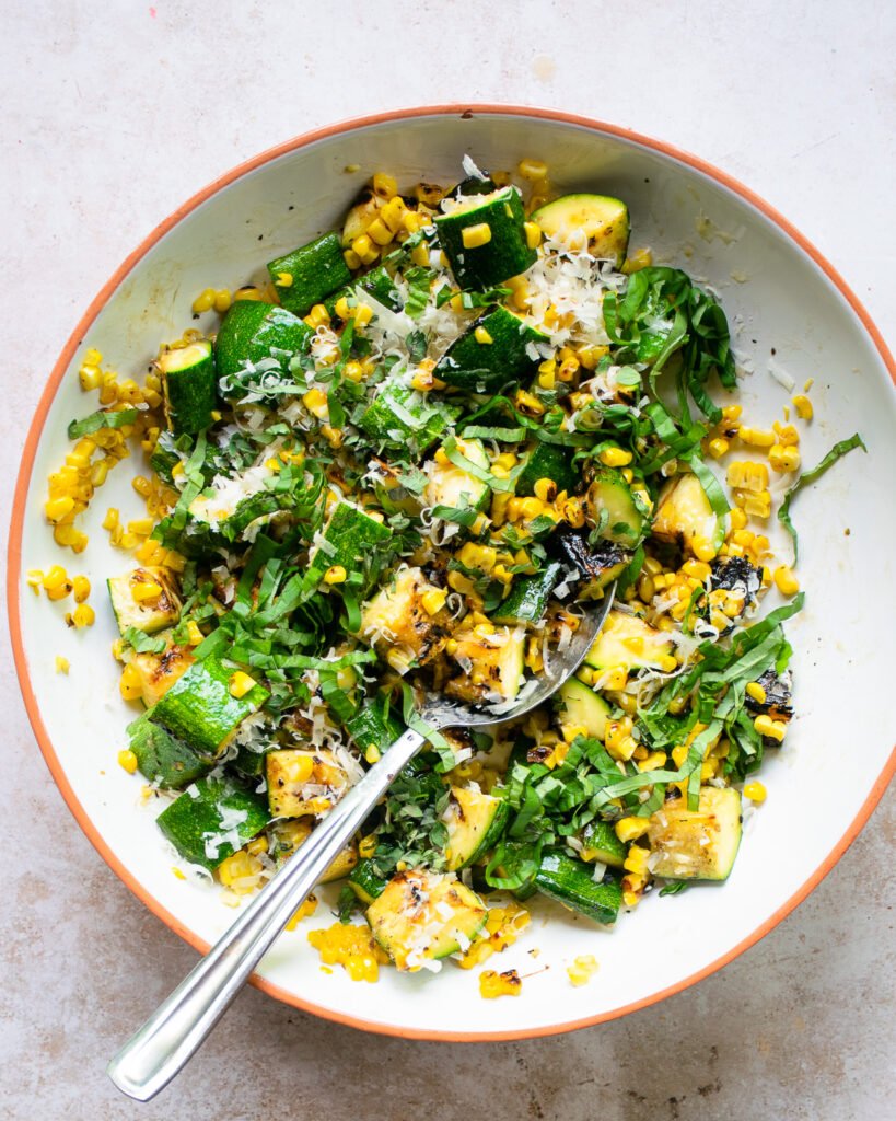 Zucchini, corn, herbs and Parmesan cheese tossed in a white bowl with a spoon.