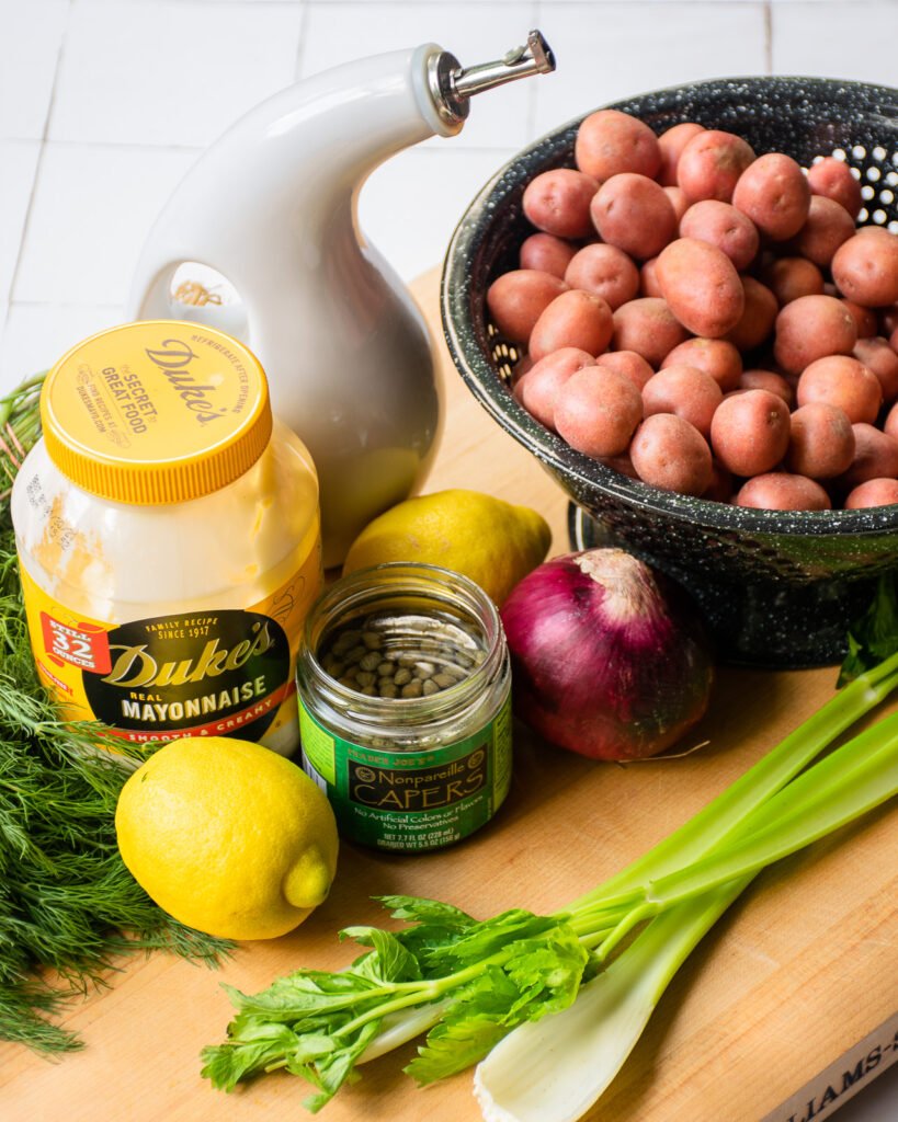 Ingredients on a cutting board - mayonnaise, lemon, capers, red onion, olive oil, celery and red potatoes.