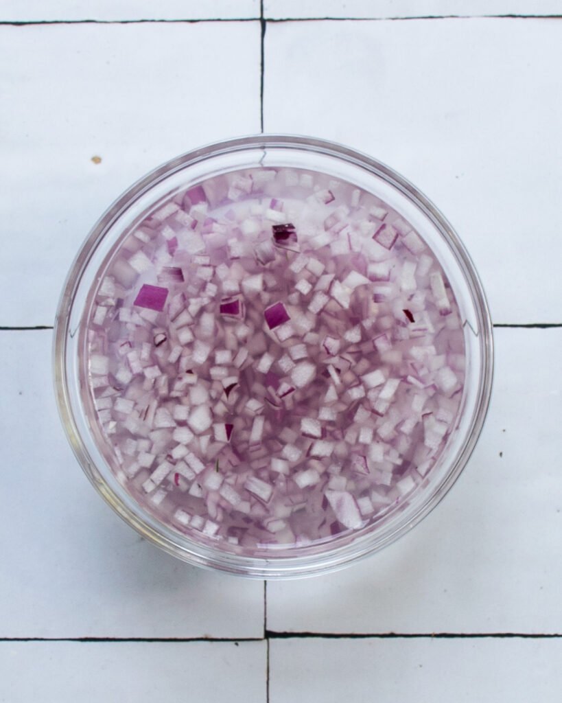 A small bowl of diced red onion in water on a tiled counter.